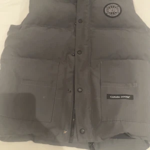 Grå dunväst från Canada Goose - Snygg grå dunväst från Canada Goose med klassisk logga på bröstet och två stora fickor framtill. Västen har dragkedja och tryckknappar, perfekt för lager-på-lager-stil under kyliga dagar.