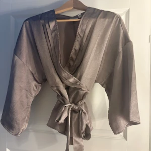 Champagnefärgad omlottopp i satin - Snygg champagnefärgad omlottopp i satin med breda trekvartsärmar och knyt i midjan. Toppens glansiga material ger en lyxig känsla och den djupa v-ringningen gör den extra trendig. Perfekt att styla med smycken för en cool look.