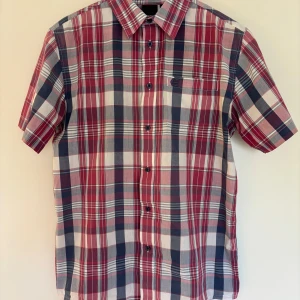 Rutig kortärmad skjorta i bomull från nederländska Etnies, storlek M - Snygg rutig skjorta i bomull från nederländska märket Etnies i vinrött, blått och vitt. Skjortan har korta ärmar, klassisk krage, knappar framtill och en bröstficka med broderad Etnies-logga. Köpt i Åre. Storlek M.