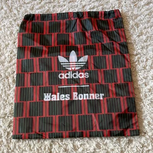 Adidas x Wales Bonner randig påse - Säljer en unik Adidas x Wales Bonner påse med rött och svart randigt mönster och gröna detaljer. Väskan har dragsko upptill och är perfekt för att bära gymkläder eller sneakers. Stor Adidas-logga och Wales Bonner-tryck på framsidan.