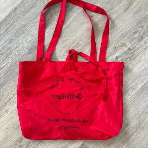 Röd shoppingväska från Valentino - Stor och rymlig röd shoppingväska från Valentino med tryckt logga och text på framsidan. Väskan har långa handtag som gör den enkel att bära på axeln. Perfekt för att ta med till stan eller skolan.