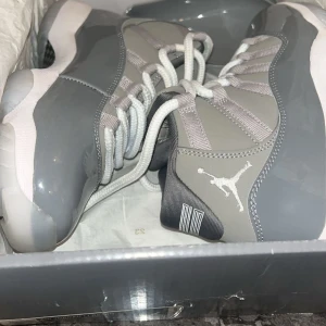 Nike Air Jordan 11 Cool Grey - Nike Air Jordan 11 Cool Grey med högt skaft, grå ovandel i mocka och lackad skinnpanel, vita snören och isblå sula. Ikonisk Jumpman-logga på hälen. Perfekta för dig som vill sticka ut med en klassisk och clean sneaker.