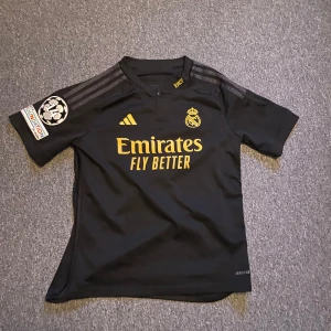 Real Madrid Champions League tröja - Real madrids bortaställ från förra säsongen med Champions league badge på armen och Vini JR på ryggen. Storlek 13-14 år. Köpt på unisport. Nypris 1250, mitt pris 300. Kan gå ner i pris vid snabb affär