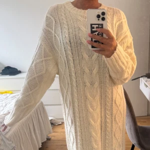 Vit stickad långklänning - Mysig vit stickad långklänning med grova kabelstickade detaljer framtill och ribbade muddar. Klänningen har lång ärm och en avslappnad, loose passform. Perfekt för dig som vill ha en chill och stilren look.