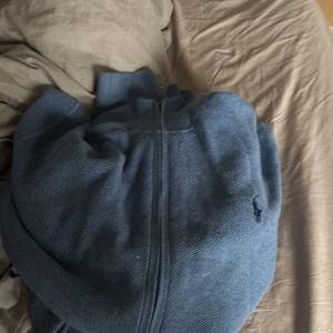 Blå kofta från Polo Ralph Lauren - Säljer en blå kofta från Polo Ralph Lauren med dragkedja framtill. Klassisk design med diskret broderad logga på bröstet. Tillverkad i mjukt stickat material som är perfekt för lager-på-lager. Passar dig som gillar stilren och tidlös look.