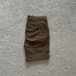 Bruna shorts från Selected Homme - Snygga bruna shorts från Selected Homme i bomull med klassisk passform. De har uppvikta benslut, bälteshällor, två sidofickor och två bakfickor med knapp. Perfekta för sommardagar och enkel att matcha med t-shirt eller skjorta. (Mest lik färgen på sista bilden). 