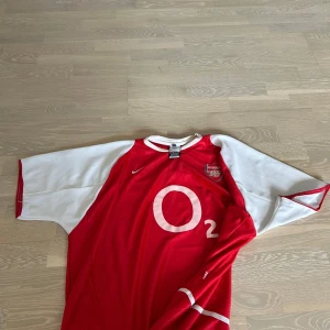 Arsenal Nike fotbollströja röd/vit - Klassisk Arsenal fotbollströja från Nike med röd kropp och vita ärmar. Stort O2-tryck på bröstet och Arsenal-emblem på vänster sida. Tröjan har rund hals och är tillverkad i lätt, ventilerande material som passar perfekt för match eller träning.