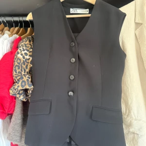 Svart väst från Zara med knappar - Stilren svart väst från Zara med klassisk v-ringning och fyra knappar framtill. Västen har två fickor med lock och ett justerbart bälte baktill för perfekt passform. Snygg och enkel design som passar till många looks.