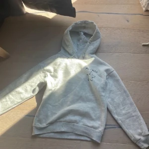 Grå hoodie med tryck 'Ma Chérie' - Säljer en ljusgrå hoodie med huva och trycket 'Ma Chérie' samt en stjärna på bröstet. Tröjan har ribbade muddar och är tillverkad i mjuk bomullsmix. Perfekt för en chill och avslappnad stil.