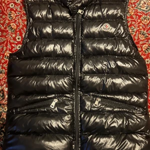 Svart dunväst från Moncler - Snygg svart dunväst från Moncler med glansig finish och klassisk logga på bröstet. Västen har två dragkedjefickor framtill och hög krage med spänne. Perfekt att slänga över en hoodie för en cool look.