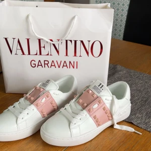 Valentino Rockstud Open sneakers vit/rosa - Snygga Valentino Rockstud Open sneakers i vitt skinn med breda rosa band och ikoniska nitar över vristen. Klassisk rund tå, platt sula och vita snören. Märkeslogga på plös och innersula. Perfekta för dig som vill sticka ut med exklusiv streetstyle.