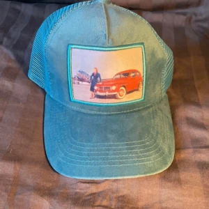 Velvet keps - Snygg turkos truckerkeps med mesh på sidorna och baksidan. Framtill sitter ett retroinspirerat tryck med en röd bil och en person, inramat i turkos. Justerbar baktill för perfekt passform. Perfekt för dig som gillar vintage vibes och coola accessoarer.
