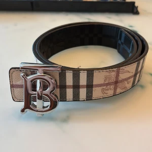 Burberry bälte - Snyggt Burberry-bälte i klassiskt rutigt mönster med beige, svart och bruna toner. Skärpet har en stor, blank silverspänne med TB-logga framtill. Tillverkat i skinn med tydlig struktur och exklusiv känsla. Perfekt för att lyfta vilken outfit som helst.  Bältet är knappt använt vi snackar bara några dagar. Prisförslag kan accepteras