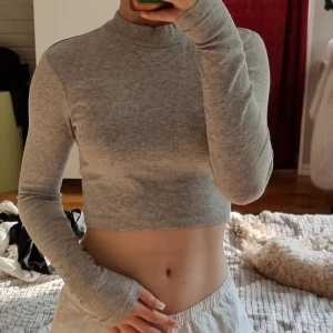 Grå croppad långärmad tröja - Säljer en ljusgrå croppad långärmad tröja med ribbad struktur. Tröjan har en hög hals och sitter tight mot kroppen, perfekt för en clean och stilren look. Passar dig som gillar minimalistiska plagg med modern passform.