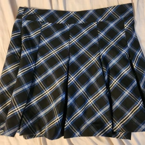 Blåsvart rutig kjol från H&M Divided - Supersnygg kort kjol i blått, svart och vitt rutigt mönster från H&M Divided. Kjolen har bred linning, veckad design och stängs med knapp och dragkedja i sidan. Perfekt för en cool och trendig look. 