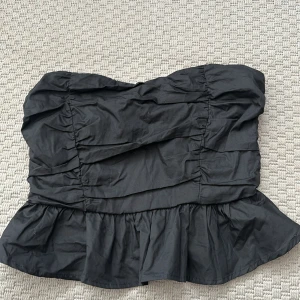 Svart volang tubtopp 36 - Snygg svart tubtopp med volangkant nertill och rynkade detaljer över hela toppen. Modellen är axelbandslös och har en trendig, lite puffig form. Storlek 36 dragkedja i ryggen 