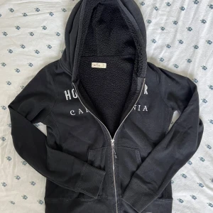 Svart hoodie från Hollister med foder - Svart vintage fodrad hoodie från Hollister med dragkedja och mysigt teddyfoder på insidan. Tröjan har huva, långa ärmar och två fickor framtill. Snyggt tryck med Hollister California på bröstet. Perfekt för kyliga dagar och chill vibes.