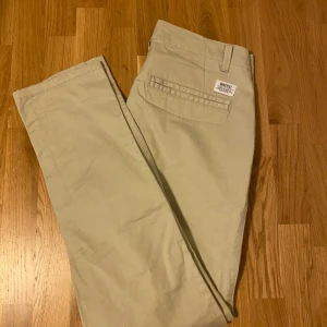 Beiga chinos från WeSC W33 L34 - Snygga beiga chinos från WeSC, modell EDU. Klassisk rak passform med diskreta bakfickor och bälteshällor. Tillverkade i bomull för en skön känsla och stilren look. Perfekta för dig som gillar enkel och clean stil.