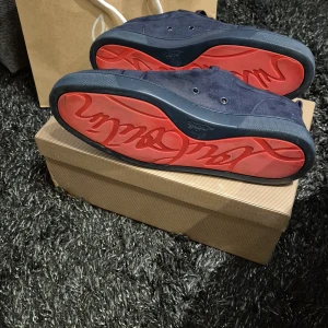 Mörkblå mockasneakers Christian Louboutin - Snygga mörkblå sneakers från Christian Louboutin i mjuk mocka med klassisk röd sula och diskret broderad logga på hälen. Skorna har rund tå, platt sula och snörning. Perfekta för dig som vill ha en lyxig och stilren look med en ikonisk touch. Den är använd ett fåtal gånger men inga defekter. Skick 8/10