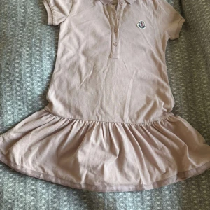 Beige kort klänning från Moncler för barn - Supersöt beige kort klänning från Moncler med pikékrage, knappar framtill och korta ärmar. Klänningen har en volang nedtill som ger en sportig men ändå gullig vibe. För barn 6år