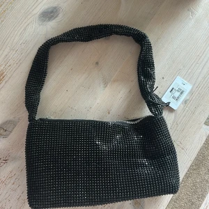 Svart glittrig axelväska från BikBok - Säljer en svart axelväska från BikBok med glittriga pärldetaljer över hela väskan. Den har en bred axelrem och stängs med dragkedja upptill. Perfekt storlek för det viktigaste och ger en snygg touch till din outfit. Inne ficka finns! Helt ny!!! 