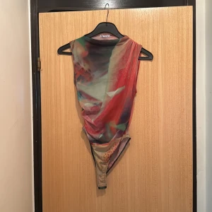 Färgglad mesh bodysuit från Gallito - Säljer en ärmlös mesh bodysuit från Gallito med abstrakt mönster i rött, grönt, beige och inslag av lila. Toppen har en tight passform och är tillverkad i tunn, stretchig polyester. Perfekt för dig som vill sticka ut med coola färger och unik design.