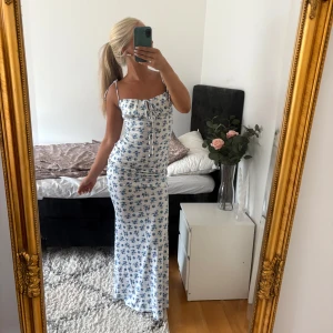 Vit långklänning med blå blommor  - Supersöt långklänning i vitt med blått blommönster utan slits. Klänningen har smala axelband som man knyter, snörning framtill och figurnära passform. Perfekt för sommarens alla tillfällen! Materialet är mjukt och stretchigt i polyester och elastan. Denna klänning är sällan använd och i fint skick! 🤍