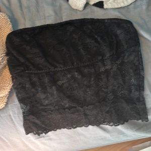 Svart spets tubtopp från SHEIN - Svart axelbandslös tubtopp från SHEIN i storlek S. Toppen är täckt av snygg spets med blommigt mönster och har en mjuk, stretchig insida. Perfekt för att skapa en cool och trendig look. Materialet är 90% polyester och 10% elastan.