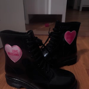 Svarta boots från Love Moschino - Coola svarta boots från Love Moschino med glansig finish och snörning. På utsidan sitter ett rosa hjärta med loggan. Insidan har ett mönster med hjärtan och Love Moschino-tryck. Perfekta för dig som vill sticka ut med en edgy vibe.
