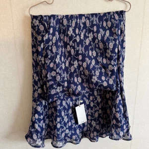 Blommig blå kjol från Mango - Supersöt blå kortkjol från Mango med blommigt mönster i vitt och ljusrosa. Kjolen har volangkant nertill och är gjord i ett lätt, flowy polyestertyg som ger en luftig känsla. Perfekt för dig som gillar romantiska vibes och vill ha något som sticker ut lite extra.