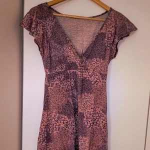 Lila blommig omlottklänning - Supersöt lila kortklänning med blommigt mönster och omlott-look. V-ringad med volangiga korta ärmar, perfekt för sommarens alla tillfällen. Lätt och luftig känsla, passar dig som gillar romantisk stil.