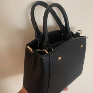 Svart handväska med gulddetaljer - LITEN svart handväska i stilren boxig modell med två handtag och guldfärgade metalldetaljer. Väskan är gjord i strukturerat skinnimitation och har plats för det viktigaste. Perfekt att bära i handen eller med axelrem. Axelrem medföljer inte