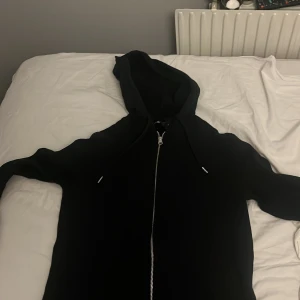 Svart hoodie med dragkedja och huva - Svart hoodie med hel dragkedja och huva. Klassisk modell med snörning i huvan och långa ärmar. Perfekt för chill dagar eller när du vill ha en enkel och stilren look.