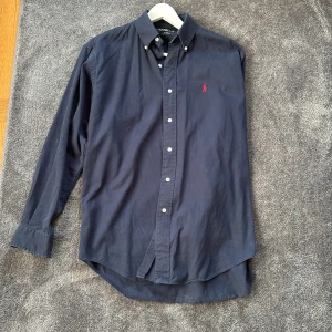 Mörkblå skjorta från Polo Ralph Lauren - Klassisk mörkblå skjorta från Polo Ralph Lauren med röd broderad logga på bröstet. Skjortan har lång ärm, knappar hela vägen och är tillverkad i 100% bomull. Perfekt för dig som gillar stilrena och tidlösa plagg. Den sitter mycket större än storlek. S sitter som en L.