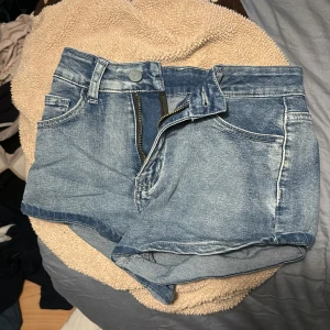 Blå jeansshorts med broderade fickor - Snygga blå jeansshorts med klassisk femficksmodell och dragkedja framtill. Baksidan har två fickor med detaljerade vita broderier som ger en unik look. Perfekta för sommaren och lätta att matcha med allt. Shortsen har bälteshällor och en något ljusare tvätt.