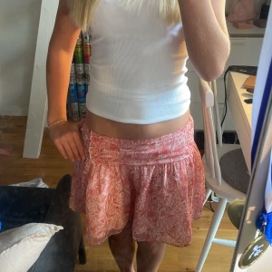 Rosa/röd  kortkjol med paisleymönster - Supersöt kortkjol i rosa/röd med paisleyinspirerat mönster. Kjolen har en lätt utställd form och bred midja, perfekt för en somrig look. Tunt, luftigt tyg med vitt foder under. Sitter snyggt på höften och är riktigt flowy. I storlek 158/164 från Ginayoung💓