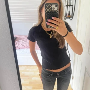 Svart ribbad topp från ONLY - Svart ribbad topp från ONLY i stretchigt material. Modellen är croppad med korta ärmar och har vågig kant nertill och vid ärmslut. Enkel och stilren, perfekt att matcha med jeans eller kjol.