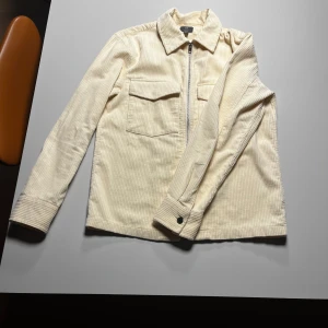 Beige manchester overshirt från H&M - Snygg beige overshirt från H&M i manchester med regular fit. Skjortan har dragkedja framtill, två bröstfickor med lock och långa ärmar med knapp vid manschetten. Perfekt att slänga över en t-shirt för en chill look.
