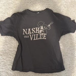 Grå Nashville t-shirt från H&M Divided - Cool grå t-shirt från H&M Divided med Nashville USA-tryck och en siluett av en gitarrspelande cowboy. Klassisk passform, rund hals och korta ärmar. Perfekt för dig som gillar musik och streetstyle. Har ett litet hål, syns på sista bilden.  Storlek XS men oversized.