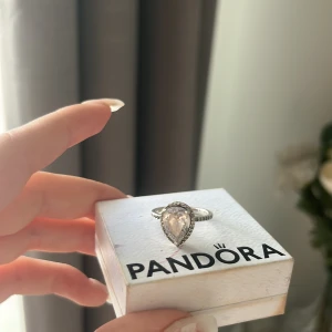 Pandora silverring med droppformad sten - Superfin silverring från Pandora med en stor, droppformad klar sten i mitten och små gnistrande stenar runt om. Ringen har en elegant och glittrig look som verkligen drar blickarna till sig. Perfekt för dig som vill ha något extra snyggt på fingret.