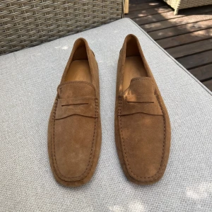 Bruna mockaloafers från Tod's - Snygga loafers från Tod's i brun mocka med klassisk pennyloafers-design. Skorna har en platt sula med svarta gummidetaljer undertill för extra grepp. Perfekta för en clean och stilren look, passar till både jeans och chinos.