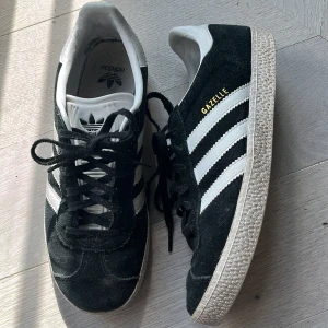 Adidas Gazelle svarta sneakers mocka - Klassiska Adidas Gazelle sneakers i svart mocka med vita detaljer och de ikoniska tre ränderna på sidorna. Skorna har snörning, platt sula och rund tå. Perfekta för en stilren och sportig look.