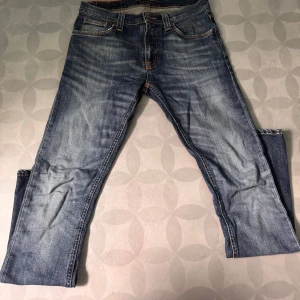 Blå jeans från Nudie Jeans Co W29 L32 - Snygga blå jeans från Nudie Jeans Co med klassisk femficksdesign och orangea sömmar. Jeansen har raka ben och coola slitningar framtill samt Nudie-logga på bakfickorna. Perfekta för en avslappnad och trendig look.