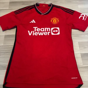 Manchester United tröja Adidas M - Röd Manchester United matchtröja från Adidas med svarta och vita detaljer på kragen och axlarna. Broderad klubbmärke, TeamViewer-tryck och Aeroready-material som andas. Slim fit-modell med korta ärmar och diskret geometriskt mönster i tyget.