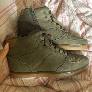 Olivgröna Isabel Marant liknande sneakers med klack - Snygga olivgröna sneakers från Diverse med högt skaft och bruna detaljer vid hälen. Skorna har snörning framtill, dragkedja på sidan och en platt, brun sula. Tillverkade i mockaliknande material som ger en cool och urban vibe.