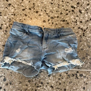 Blå jeansshorts med slitningar &DENIM - Snygga blå jeansshorts från &DENIM med slitna detaljer och fransig kant nertill. Shortsen har klassisk femficksmodell och coola trasiga partier framtill. Perfekta för en avslappnad sommarstil.