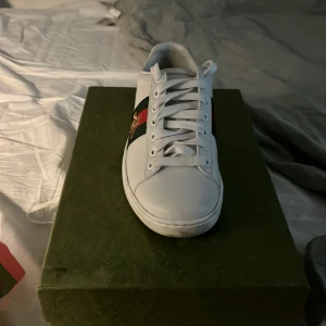 Gucci Ace vita sneakers med bi-detalj - Gucci Ace sneakers i vitt skinn med klassiska gröna och röda ränder på sidan och broderad guldfärgad bi. Baksidan har metallicröd och metallicgrön häl med ormmönster och Gucci-logga. Platt sula och snörning framtill. Riktigt ikoniska och snygga sneakers.
