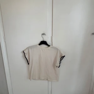 Vit topp med mörkblåa volangdetaljer - Säljer en söt vit topp med korta ärmar och mörkblåa volangdetaljer längs ärmsluten. Toppen har en rak och lite loose passform, perfekt till jeans eller kjol.