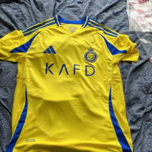 Al Nassr Ronaldo 7 matchtröja Adidas XL - Gul och blå Al Nassr fotbollströja från Adidas i storlek XL, slim fit. Tröjan har korta ärmar, klubbmärke på bröstet, Ronaldo 7-tryck på ryggen och Aeroready-material som andas. Perfekt för dig som vill supporta Ronaldo och Al Nassr på riktigt. Storleken är XL passar dom som är 180-185 cm lång. Vid minsta fundering eller om du vill ha mer bilder på tröjan hör av dig.