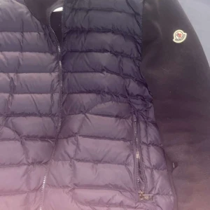Svart Moncler dunjacka med dragkedja - Säljer en svart dunjacka från Moncler med quiltad framsida och släta ärmar. Jackan har tydliga dragkedjor och klassisk Moncler-logga på ärmen. Perfekt för dig som vill ha en stilren och varm jacka med snygga detaljer.